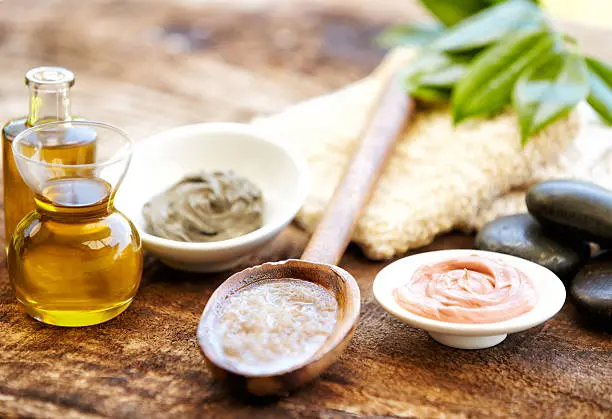 Organic mud mask with moisturizer, massage oil, washcloth, green leaves and salt scrub on a wooden spoon. Everything is set on wooden distressed surface outdoors. Horizontal shot.